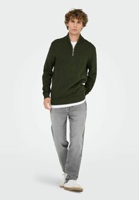 Dunkelgrüner Strickpullover mit Fischgrätenmuster, halber Reißverschluss-Kragen, gerippte Bündchen, über einem weißen T-Shirt getragen, kombiniert mit grauen Jeans.