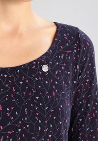 Mörkblå topp med rund halsringning, prydd med ett blommönster i rosa och blå nyanser. Har en silverfärgad knappdetalj vid halsringningen.