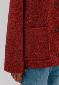 Rote strukturierte Jacke mit einer flauschigen Oberfläche, mit einer aufgenähten Tasche und schwarzen Knöpfen. Schulter- und Ärmeldetails sind im Kontrast zu Denim sichtbar.