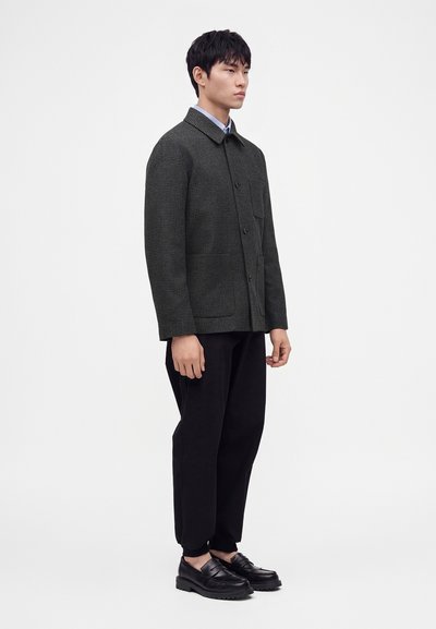 Homme portant une veste à carreaux gris foncé, un pantalon noir et des mocassins en cuir noirs, debout devant un fond clair uni.