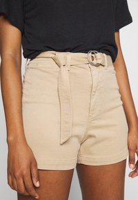 Woman wearing beige high-waisted shorts with a fabric belt and black tucked-in t-shirt, hands relaxed by her sides.