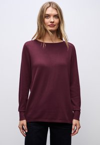 Bordeaux lange mouw gebreid top met een boatneck en geribde manchetten, gecombineerd met donkere jeans tegen een neutrale achtergrond.