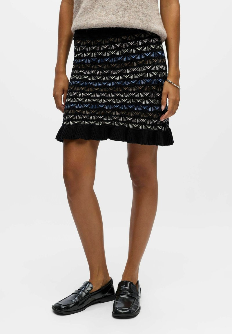 Jupe noire en tricot avec un motif bleu et blanc, ornée d'un ourlet volants. Assortie à des mocassins noirs brillants.