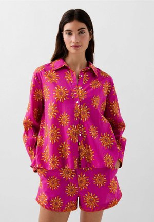 Camisa rosa de corte amplio con botones y patrones de sol en naranja y amarillo, combinada con pantalones cortos a juego. Presenta cuello y bolsillos laterales.