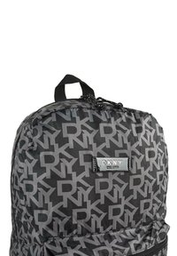 Mochila negra de nailon con un patrón de logotipos grises repetidos, placa de marca brillante y un asa superior para llevar. Incluye un bolsillo frontal con cremallera.