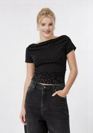 Νεαρή γυναίκα με ξανθά μαλλιά σε αλογοουρά που χαμογελά, φοράει μαύρο τοπ με λευκά πουά και τζιν μαύρο ψηλόμεσο, με το χέρι στην τσέπη.