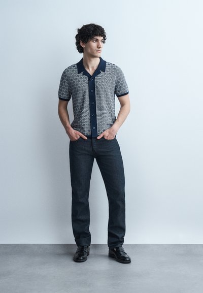 Jeune homme aux cheveux bouclés se tenant contre un mur blanc, portant une chemise à manches courtes boutonnée à motifs, un jean foncé et des chaussures noires.