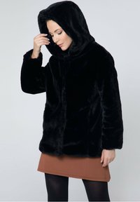Čierny umelý kožuch s kapucňou, ktorý má jemnú textúru a predné zapínanie, kombinovaný s hnedou sukňou a čiernymi pančuchami.