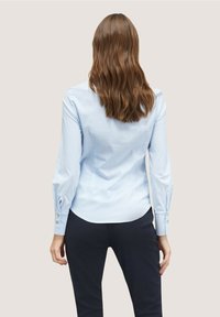 Camicia button-up a righe blu chiaro con maniche lunghe, orlo curvo e polsini abbottonati, abbinata a pantaloni scuri su misura.