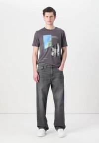 Grå grafisk t-skjorte med abstrakte mønstre og tekst, kombinert med løse mørke jeans. Modellen har på seg hvite joggesko mot en nøytral bakgrunn.