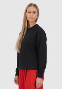 Felpa nera con colletto rotondo e maniche raglan, realizzata in un tessuto morbido; presenta polsini a costine. Abbinata a pantaloni rossi.