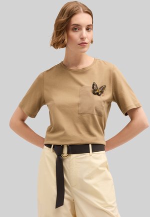 Bruine katoenen t-shirt met korte mouwen, voorzien van een borstzak en een decoratieve vlinder-speld. Gecombineerd met hooggecapte lichte broek.