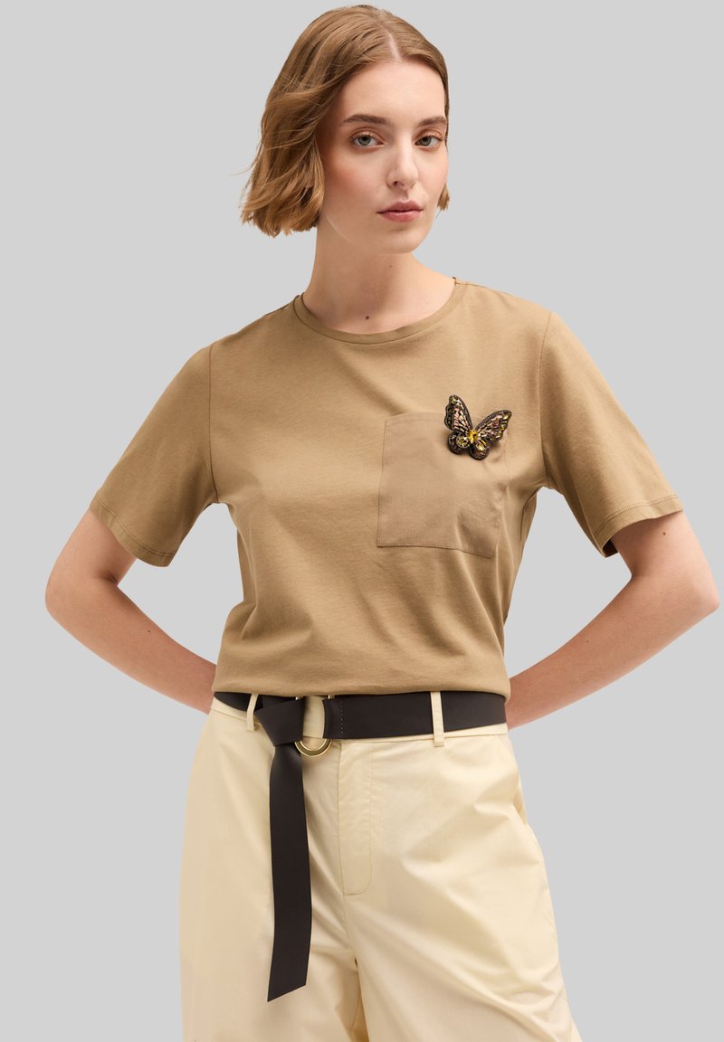 Camiseta de algodón marrón con mangas cortas, con un bolsillo en el pecho y un broche decorativo en forma de mariposa. Combinada con pantalones claros de cintura alta.