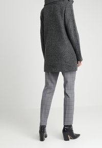 Grijze wolmix jas met een hoge kraag, gecombineerd met grijze geruite broeken en zwarte enkellaarsjes met hak. Glad textuur, nauwsluitende pasvorm.