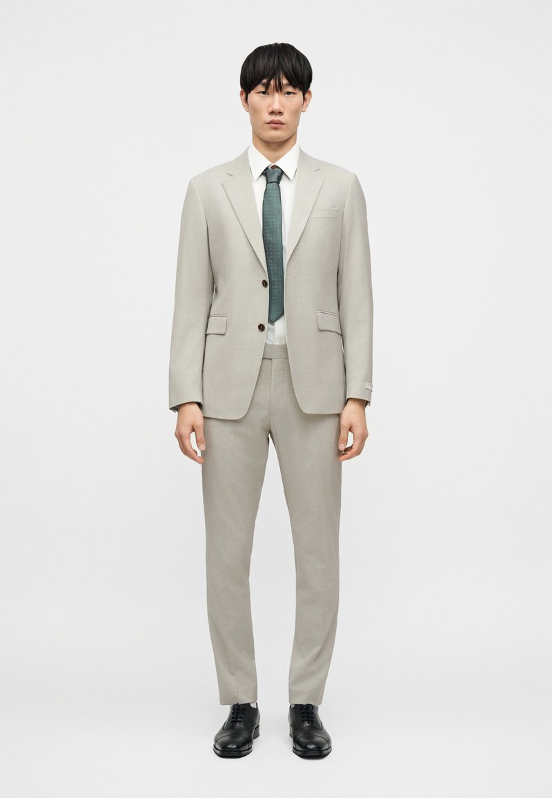 Ανοιχτό γκρι κοστούμι δύο κομματιών με εφαρμοστή σακάκι και ταιριαστό παντελόνι. Περιλαμβάνει λευκό πουκάμισο και πράσινη γραβάτα. Μαύρα επίσημα παπούτσια ολοκληρώνουν την εμφάνιση.