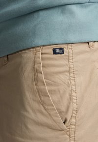 Beige katoenen broeken met kenmerkende zijzakstiksel, met een blauwe label "Petrol Industries" op de tailleband.