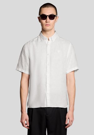 Mann trägt schwarze Sonnenbrille, weißes Kurzarm-Hemd mit Knöpfen und Kragen sowie schwarze Hose, steht vor einem schlichten grauen Hintergrund.