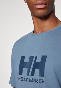 Zilā kokvilnas T-kreklā ar lielu tumši zilā krāsā "HH" logotipu un "HELLY HANSEN" tekstu, ar apaļu apkakli un īsām piedurknēm.