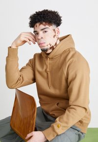Brown hoodie with a drawstring, seated against a light background, holding a wood-like chair. Features a distinct fabric texture and logo detail.