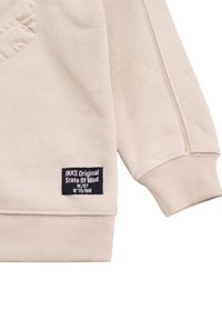 Felpa di un beige chiaro con polsini e orlo a costine, caratterizzata da un logo in rilievo e un'etichetta nera che riporta "IKKS Original State Of Mind."