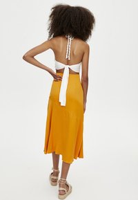 Vestido midi amarillo con una silueta fluida, que presenta una tirante halter atado en blanco y una textura suave, combinado con sandalias trenzadas.