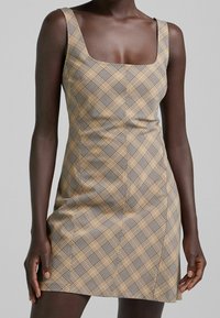 Beige rutigt miniklänning med fyrkantig halsringning, breda axelremmar och vertikala sömmar. Mönstret har svart och vit rutig detaljer.