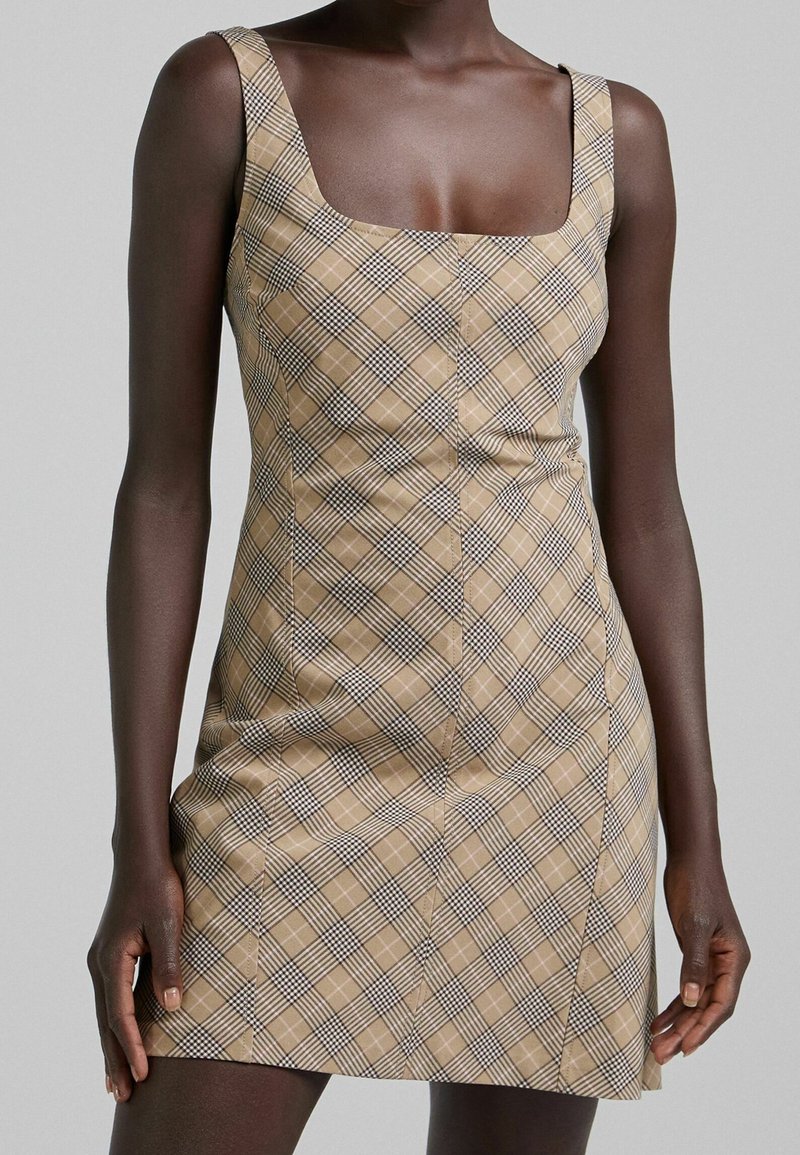 Beige rutigt miniklänning med fyrkantig halsringning, breda axelremmar och vertikala sömmar. Mönstret har svart och vit rutig detaljer.