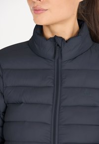 Mörk vadderad jacka med hög krage, framzipper och horisontell sömnad. Materialet verkar slätt och lätt, med en subtil logotyp.
