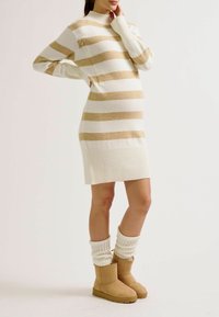 Robe pull rayée beige et blanche avec ourlet côtelé, col montant et détail fermeture éclair latérale, portée avec des bottines camel et des guêtres crème.