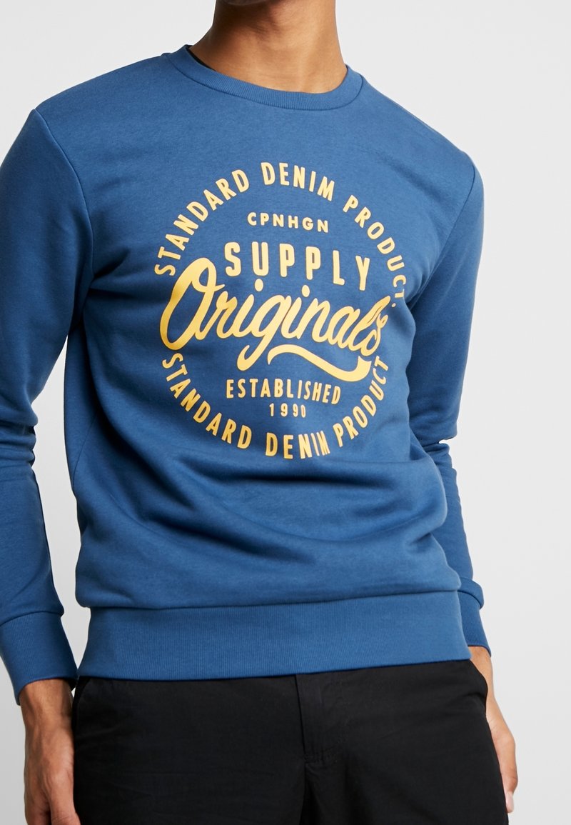 Blå sweatshirt med rund hals, långa ärmar och en stor gul grafisk textdesign med "Supply Originals" och "Standard Denim."
