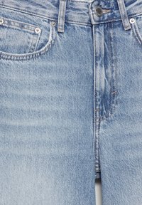 Jeans en denim bleu clair avec bouton devant, fermeture éclair, passants de ceinture et coutures visibles autour des poches et des coutures.