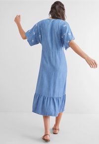 Jeanskleid in Hellblau mit kurzen, lockeren Ärmeln, rüschigem Saum und floralen Stickdetails auf den Schultern. Flache Sandalen sind sichtbar.