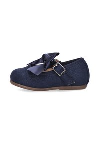 Zapato plano de tela con purpurina azul marino y un lazo ancho de satén en la parte delantera. Presenta una correa con hebilla y una suela flexible de color marrón.