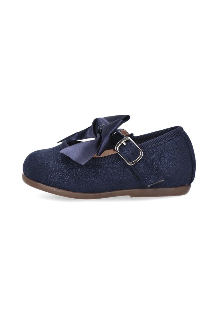 Zapato plano de tela con purpurina azul marino y un lazo ancho de satén en la parte delantera. Presenta una correa con hebilla y una suela flexible de color marrón.