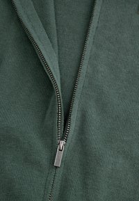 Zielony rozpinany ubiór o drobno dzianym wzorze. Posiada metalowy zamek błyskawiczny z prostokątnym uchwytem, odsłaniający klasyczny kołnierzyk.