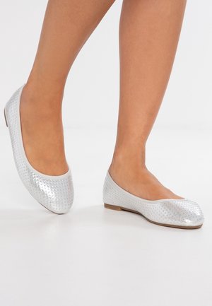 Beine mit silbernen Ballettschuhen mit kleinen Strasssteinen, stehend auf einem weißen Boden vor einem schlichten hellen Hintergrund.
