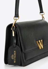 Borsa a mano in pelle nera con finitura testurizzata, dettagli in oro e un prominente logo dorato a forma di "W". Presenta un'etichetta removibile con la scritta "Wittchen".