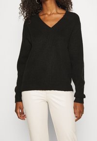 Schwarzer Strickpullover mit V-Ausschnitt und langen Ärmeln, aus strukturiertem Stoff mit gerippten Bündchen, kombiniert mit hellfarbigen Hosen.