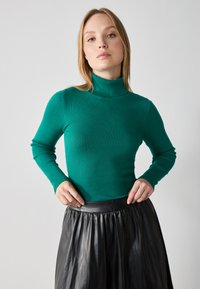 Turtleneck genser i ribbet grønn stoff, tettsittende design med lange ermer, kombinert med et svart plisse lærsnor i midjen.