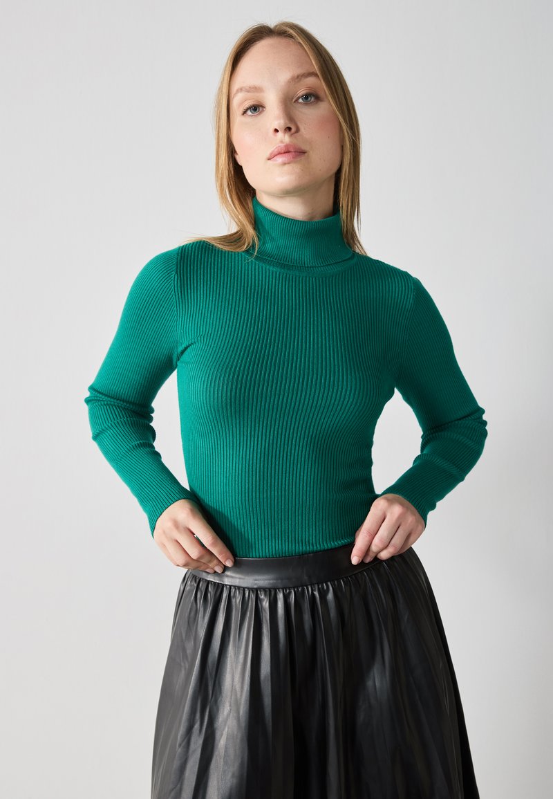 Turtleneck genser i ribbet grønn stoff, tettsittende design med lange ermer, kombinert med et svart plisse lærsnor i midjen.