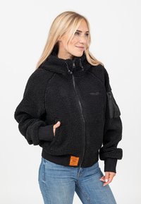 Veste en polaire noire avec un col montant, fermeture éclair, poches latérales et une poche sur la manche avec fermeture éclair. Comprend une étiquette de marque orange.