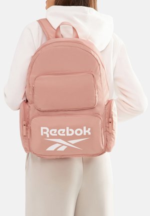 Mujer con sudadera blanca con capucha y pantalones claros, con una mochila rosa de Reebok que tiene múltiples compartimentos con cremallera.