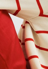 Strickpullover aus cremefarbigem Stoff mit roten Streifen, drapiert über einem einfarbigen roten Textil. Die Textur erscheint weich und leicht strukturiert.