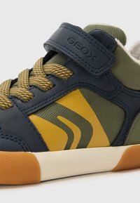 Blå og grøn sneaker med gule accenter, udstyret med en tekstureret overdel, mønstret sidesektion og en velcrolukning.