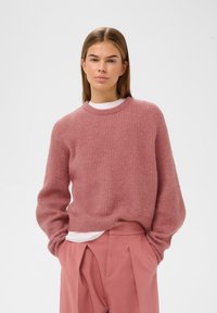 Lyserød strikket sweater med ribbet tekstur og rund halsudskæring, lagdelt over en hvid skjorte, båret med matchende lyserøde højprofilerede bukser.