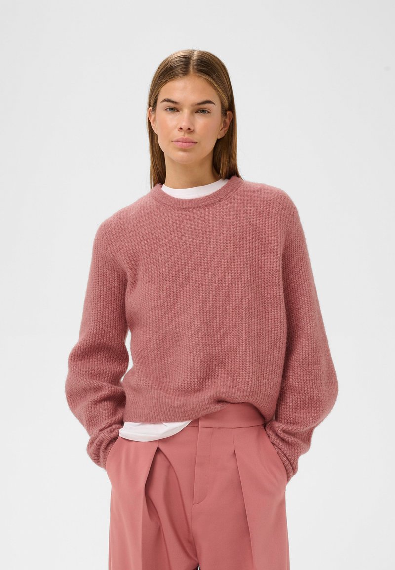 Lyserød strikket sweater med ribbet tekstur og rund halsudskæring, lagdelt over en hvid skjorte, båret med matchende lyserøde højprofilerede bukser.