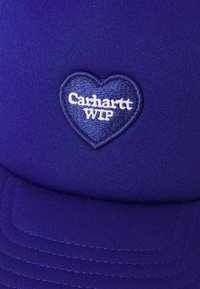 Lila Kappe mit glattem Stofffinish, versehen mit einem herzförmigen Patch und weißem gesticktem Text "Carhartt WIP." Genähte Details auf dem Schirm.