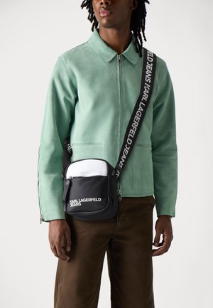 Grüne Wildlederjacke mit kragenschnitt, kombiniert mit einer schwarz-weißen Schultertasche mit Logo und verstellbarem Riemen.