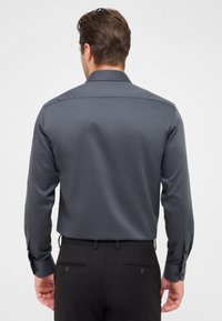 Maglietta grigia scuro a maniche lunghe con una texture liscia, colletto a punta e polsini con bottoni, abbinata a pantaloni neri con tasche posteriori.