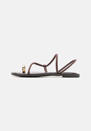 Flache schwarze Sandale mit dünnen braunen Riemen, einem goldenen Zehenring-Detail und Slingback-Stil vor einem hellgrauen Hintergrund.